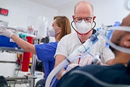 Ein Arzt in der zentralen Notaufnahme versorgt einen Patienten und wird von einer Kollegin im Hintergrund unterstützt.