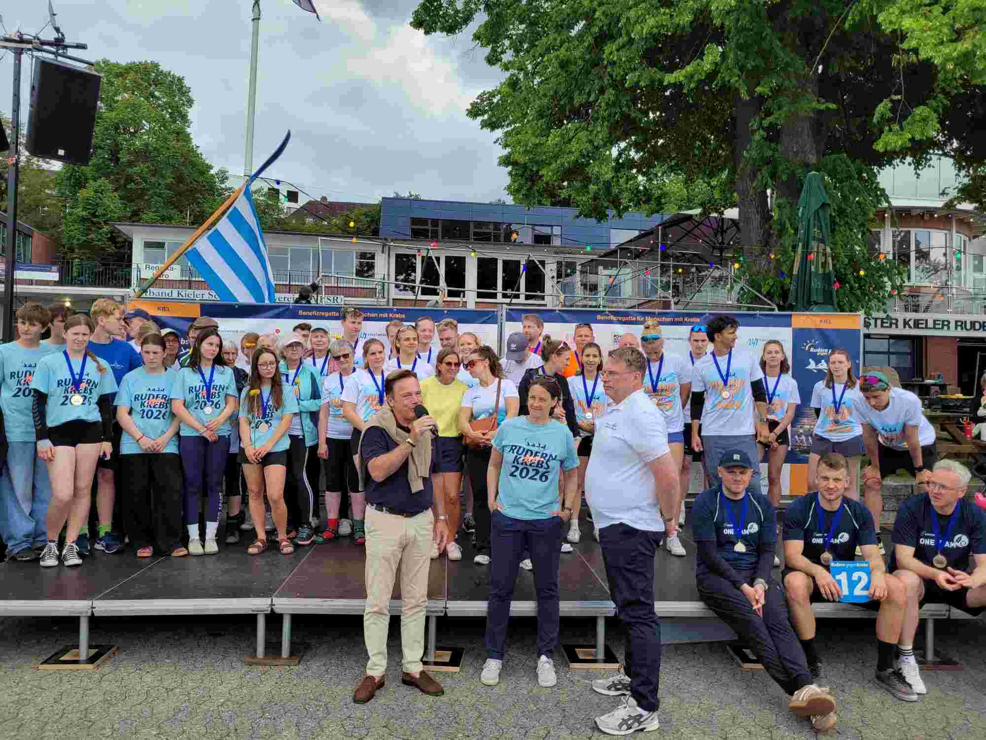 Benefizregatta Rudern gegen Krebs. Im Hintergrund alle Teilnehmer. Im Vordergrund: Gewinner der Kategorien Einsteiger, Könner mit Patienten- und Schülerteams sowie alle ehrenamtlichen Helfer*innen, vorne Moderator Andreas Otto mit Oliver Bettzieche, Stiftung Leben mit Krebs und Maja Darmstadt vom VKSR