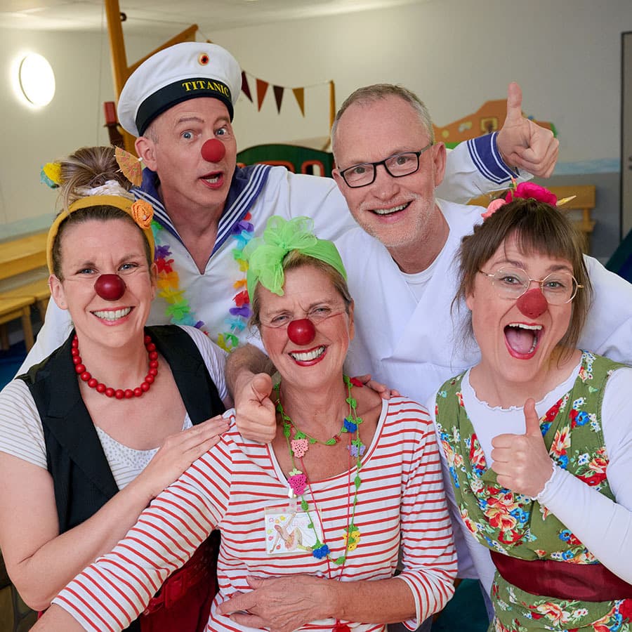 Vier kostümierte Klinikclowns stehen zusammen mit dem Chefarzt der Kinderklinik Herrn Doktor Ankermann und lachen in die Kamera.