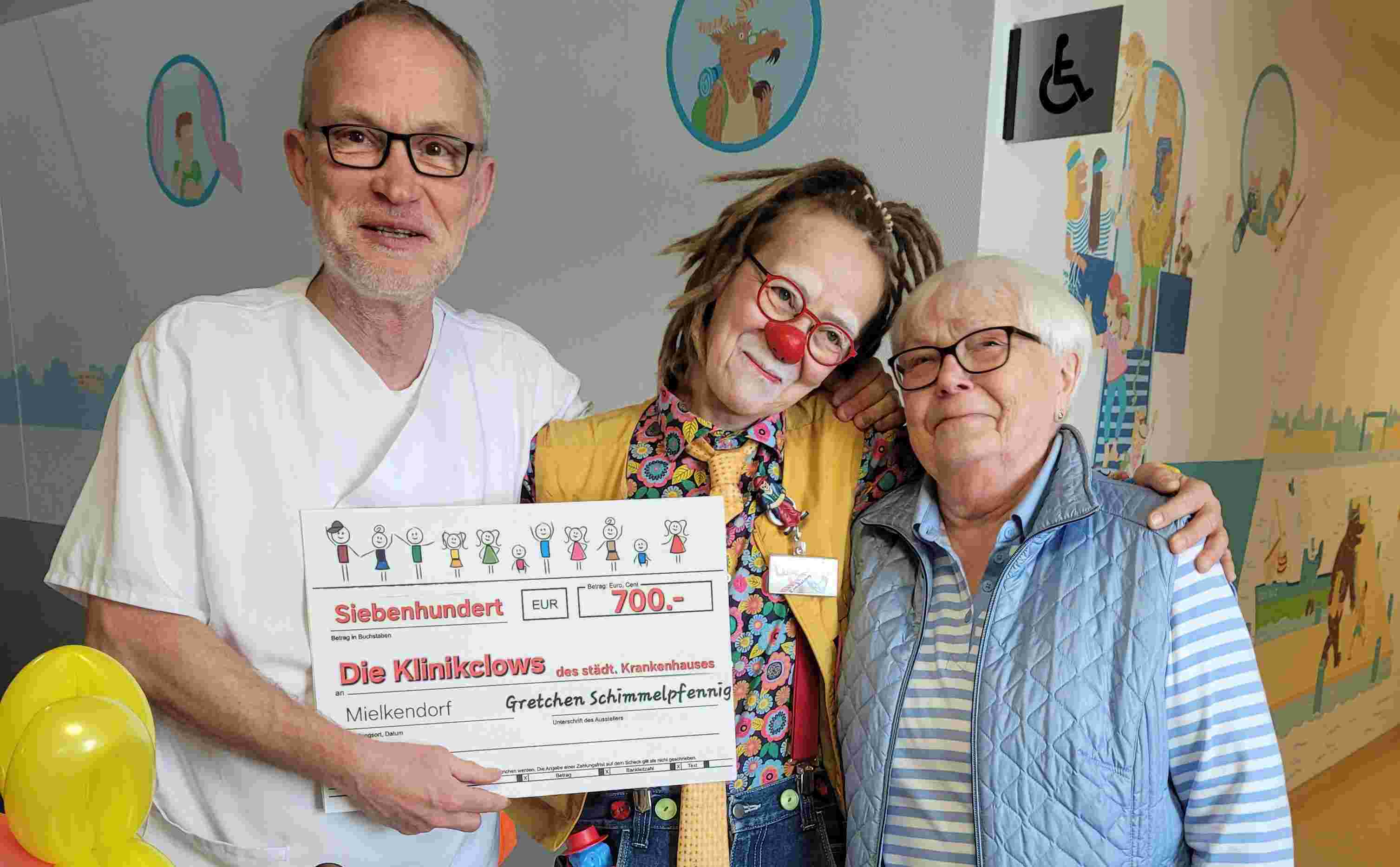 Empfang der Spendenübergabe. Chefarzt Dr. Ankermann nimmt den Scheck von Frau Grete Schimmelpfenning im Empfang. In der Mitte ist Klinikclownin "Upps" zu sehen.