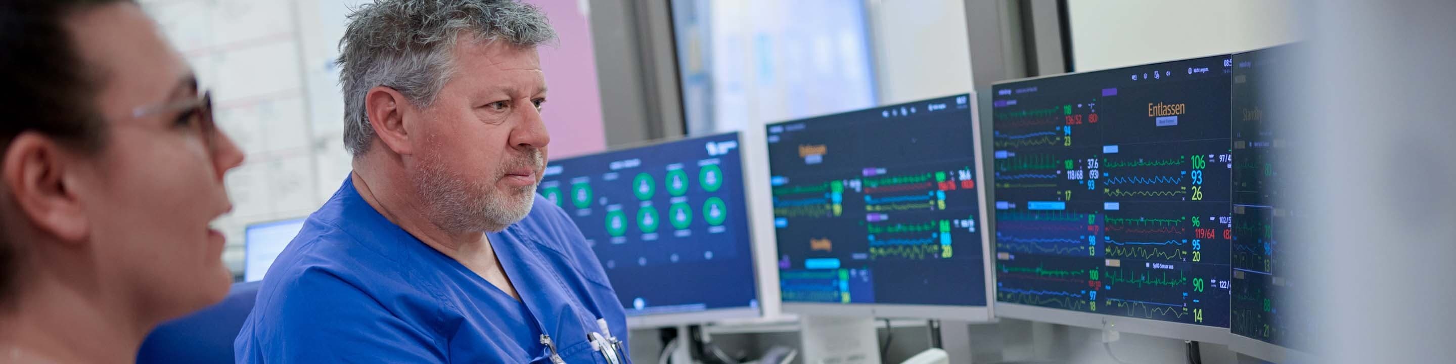 Eine Kollegin und ein Kollege der Intensivstation sitzen in einem Beobachtungsraum vor zahlreichen Bildschirmen und überprüfen medizinische Werte von Patiente*innen.
