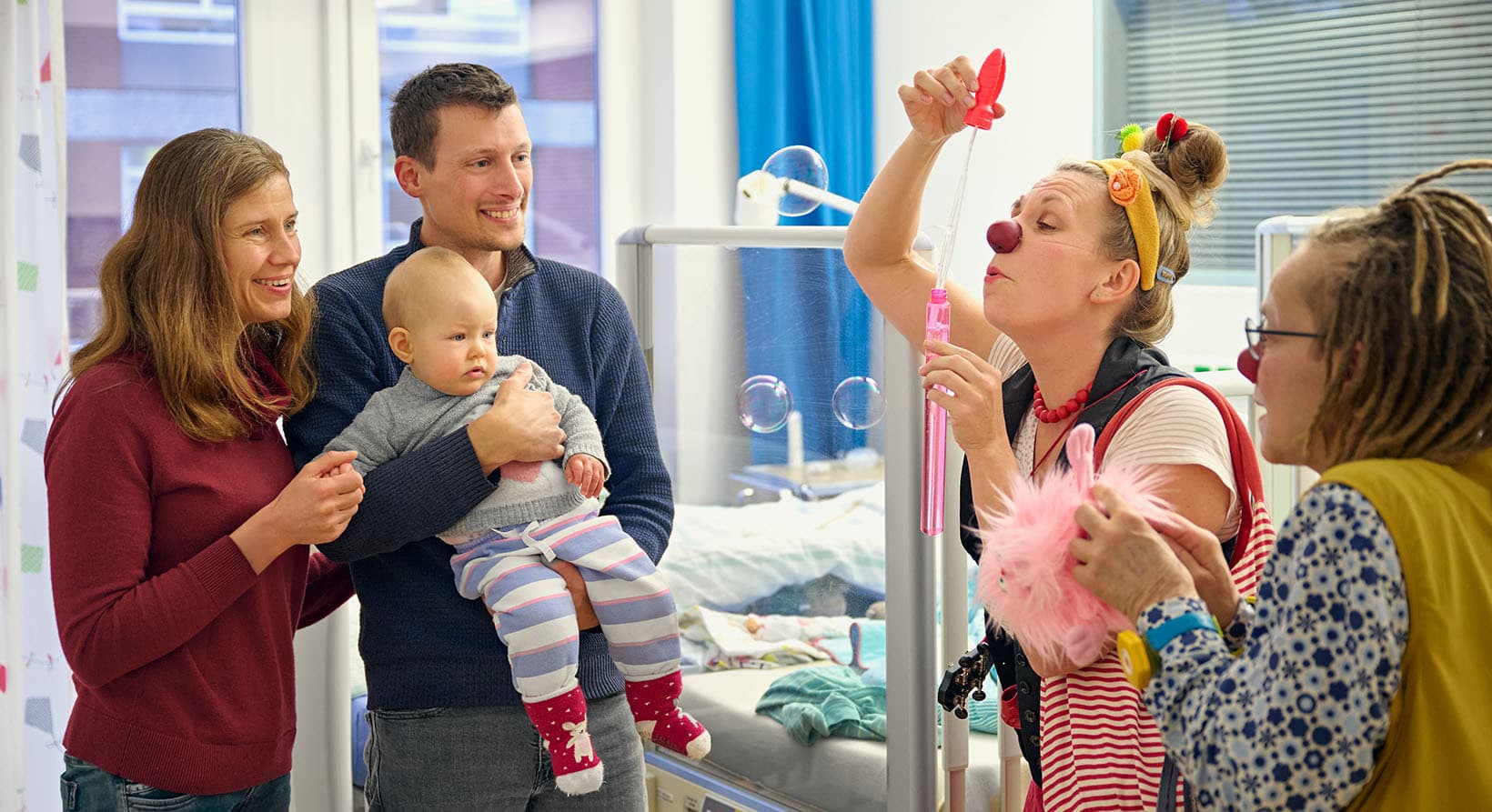 In einem Patientenzimmer schauen sich Eltern lachend mit ihrem Kind auf dem Arm, zwei Klinikclowns in Aktion an.