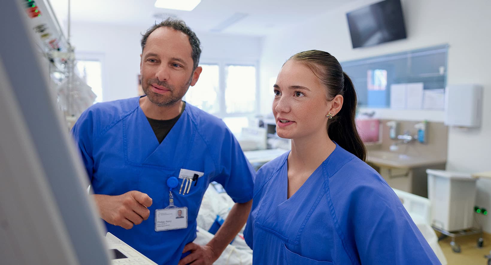 Eine Kollegin und ein Kollegin der Intensivstation stehen zusammen in einem Patientenzimmer und beobachten die Werte eines Patienten auf einem Monitor.