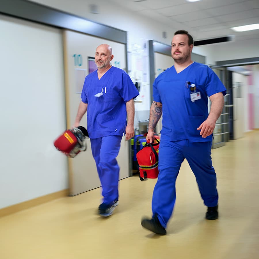 Zwei Mitarbeiter der Intensivstation laufen zusammen mit einer medizinischen Notfallausrüstung über einen Flur.