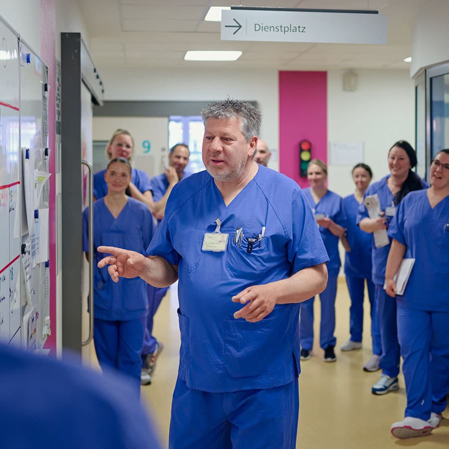 Das Team der Intensivstation steht auf dem Flur des Bereichs vor einer großen Tafel, um alle Patient*innen zu besprechen und gemeinsam Benhandlungsziele festzulegen.