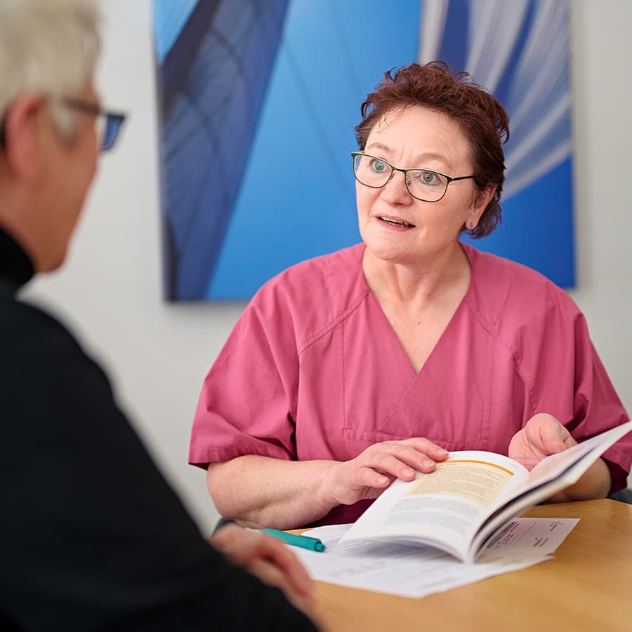 Eine Mitarbeiterin mit einer Krankenhausbroschüre in der Hand im Gespräch mit einer Patienten.
